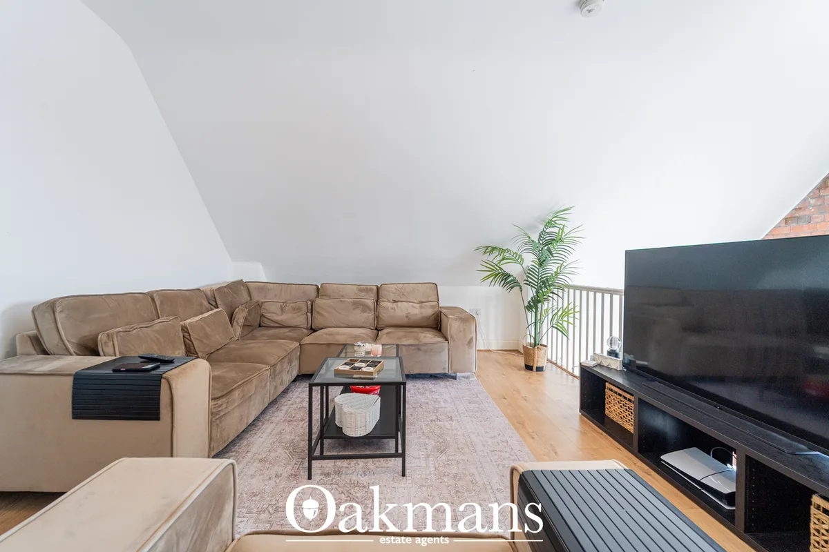 Open-plan living room with beige sectional sofa, black TV unit, wooden flooring, potted plant, and sloped ceiling with s