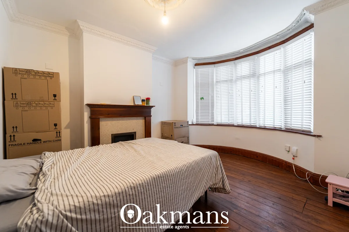 Spacious bedroom with striped bedding, ornate fireplace, bay window with white shutters, hardwood flooring and cardboard