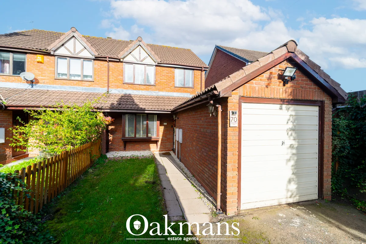 Red brick semi-detached house with white garage door, pitched roof, front garden with green fence and mature shrubs