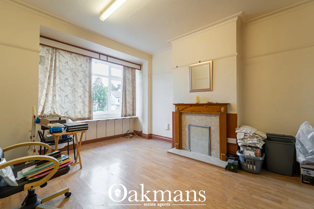 Spacious living room with wooden fireplace, large bay window with patterned curtains, wooden flooring, and cream walls