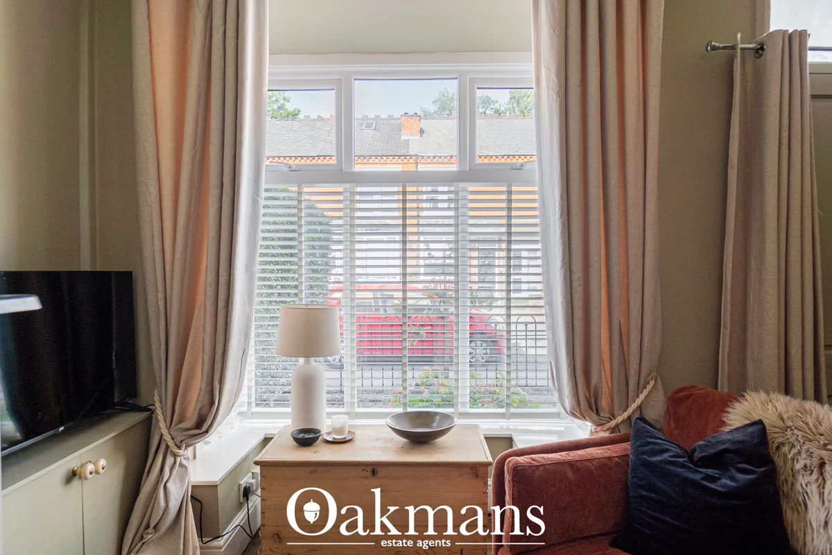 Bright living room with large multi-pane window, striped blinds, beige curtains, burgundy sofa with cushions, wooden sid