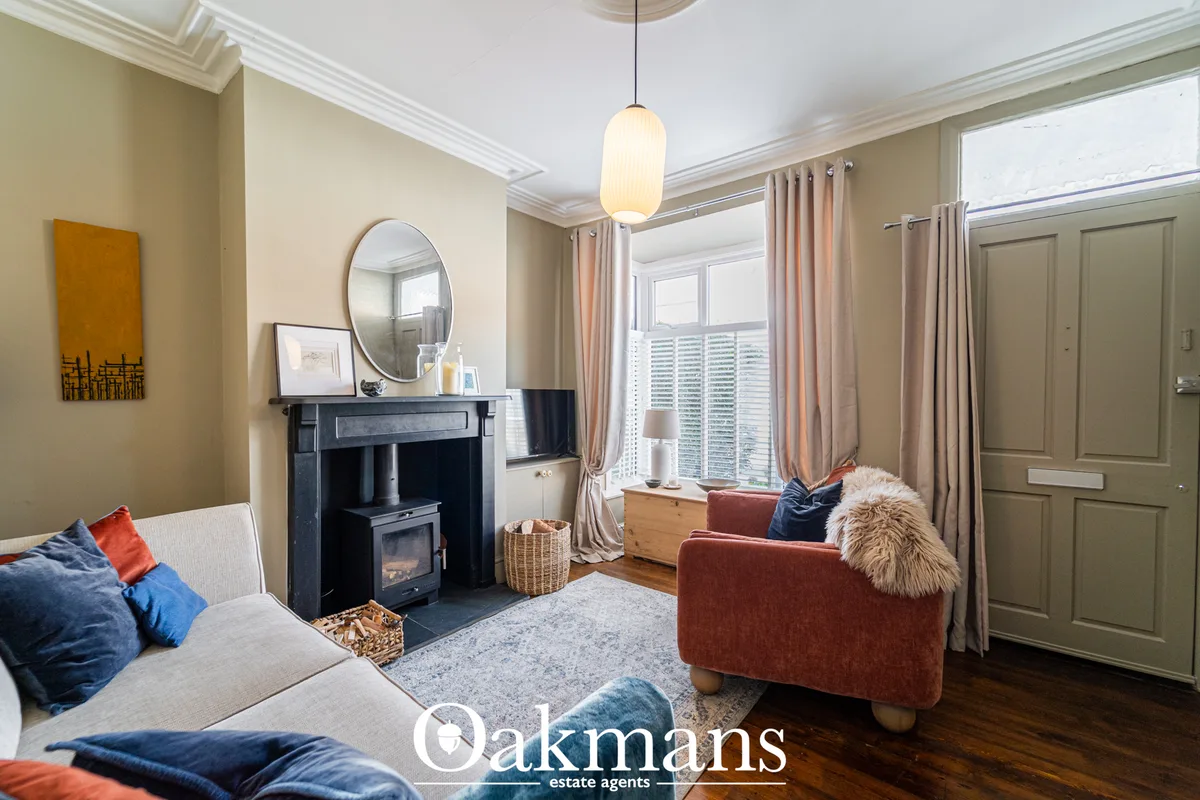 Bright living room with cream walls, black wood stove, bay window with dual curtains, gray sofa with blue and rust cushi