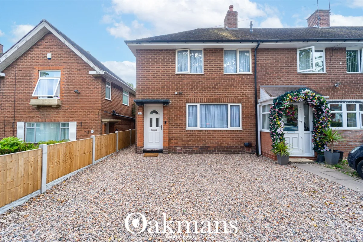 Red brick semi-detached house with white door, gravel driveway, wooden fence and decorative flowering arch entrance