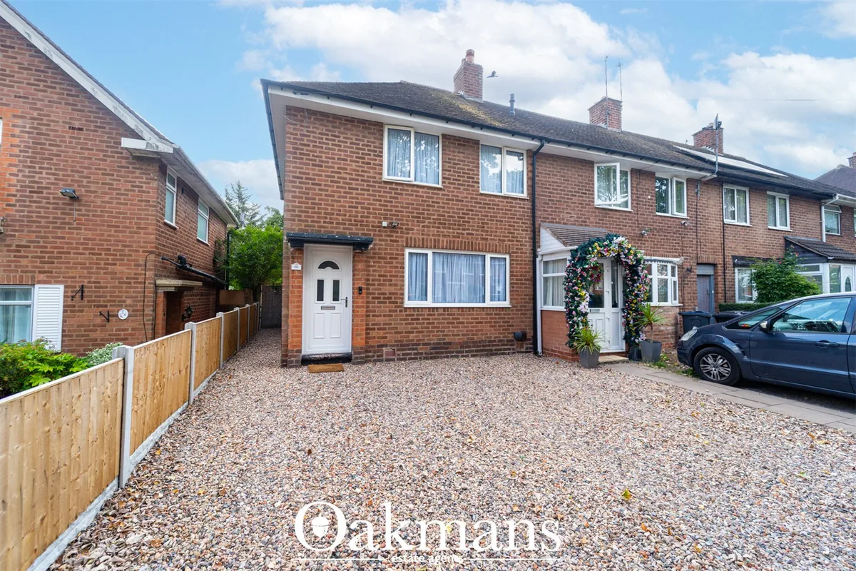Red brick semi-detached house with white door, gravel driveway, wooden fence, parked car, and flowering entrance arch