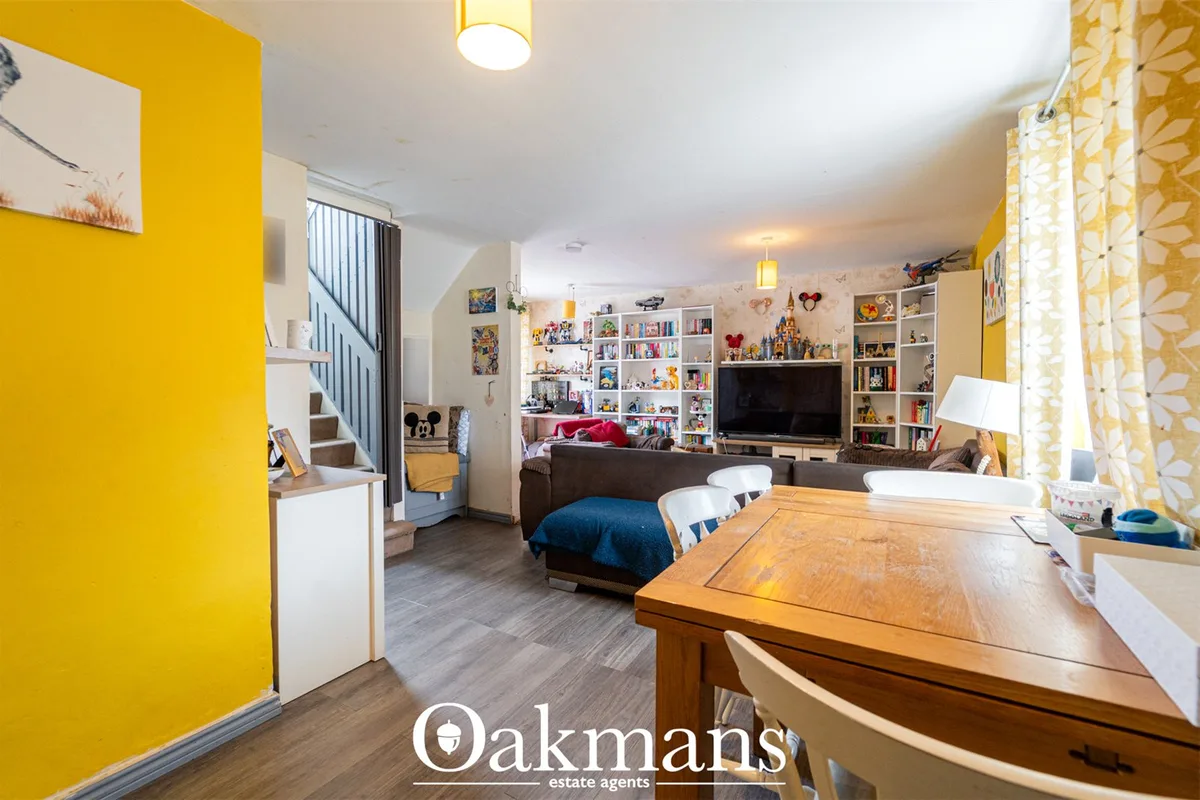 Open-plan living space with yellow feature wall, wooden dining table, brown sofa, TV, shelving with collectibles, stairc