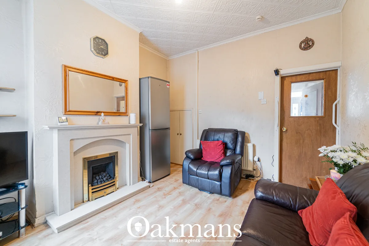 Living room with fireplace, cream walls, dark leather sofa, wooden door, kitchen visible in background