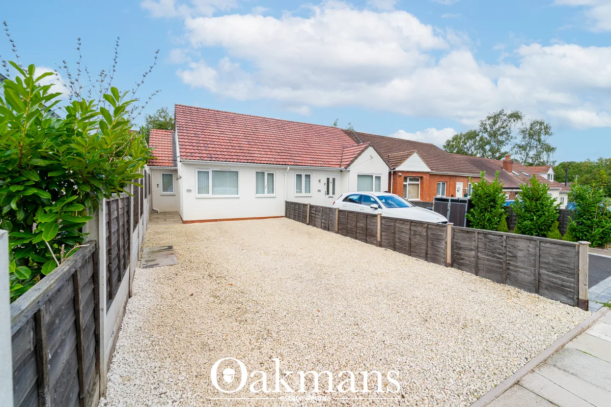 White bungalow with red tiled roof, gravel driveway, parked car, wooden fencing, green plants and clear sky