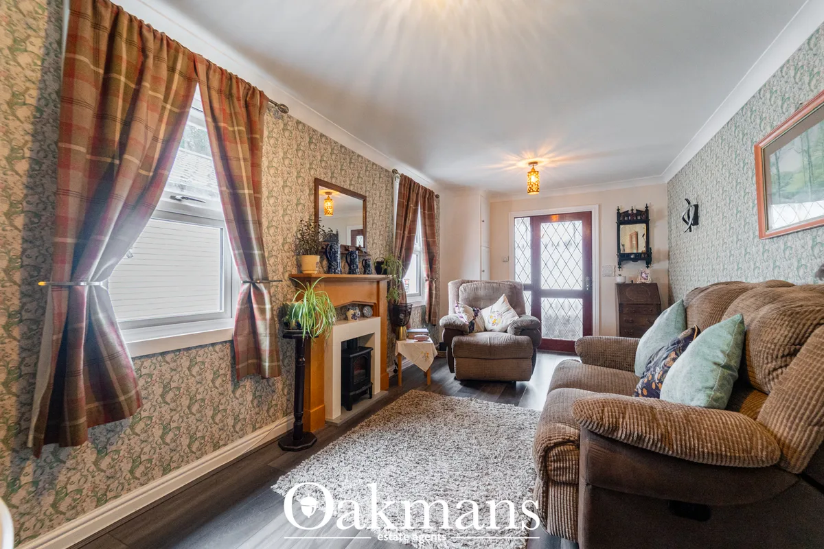 Spacious living room with beige patterned wallpaper, comfortable upholstered seating, traditional fireplace, plaid curta