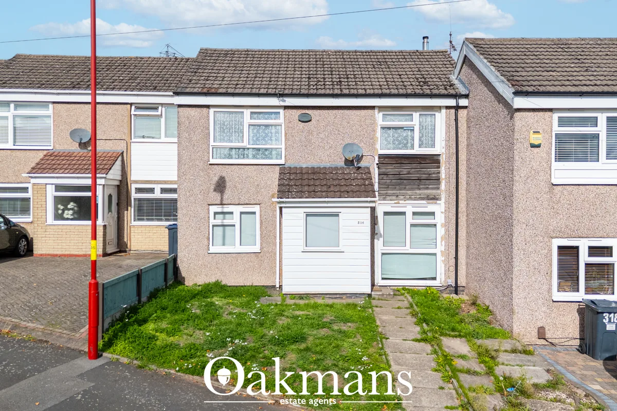 Semi-detached brick house with white garage door, overgrown front garden, parked cars and street parking visible