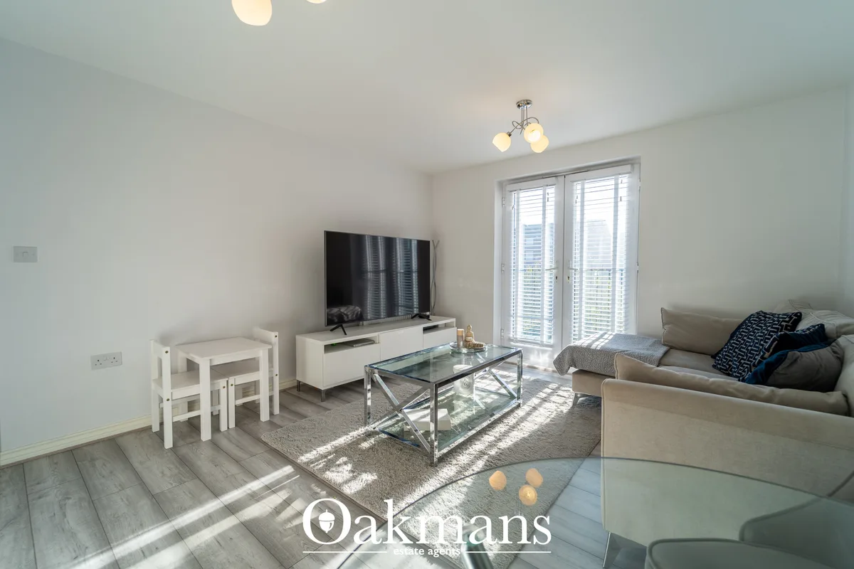 Bright living room with beige sofa, glass coffee table, white TV unit, wooden flooring, and French doors opening to balc