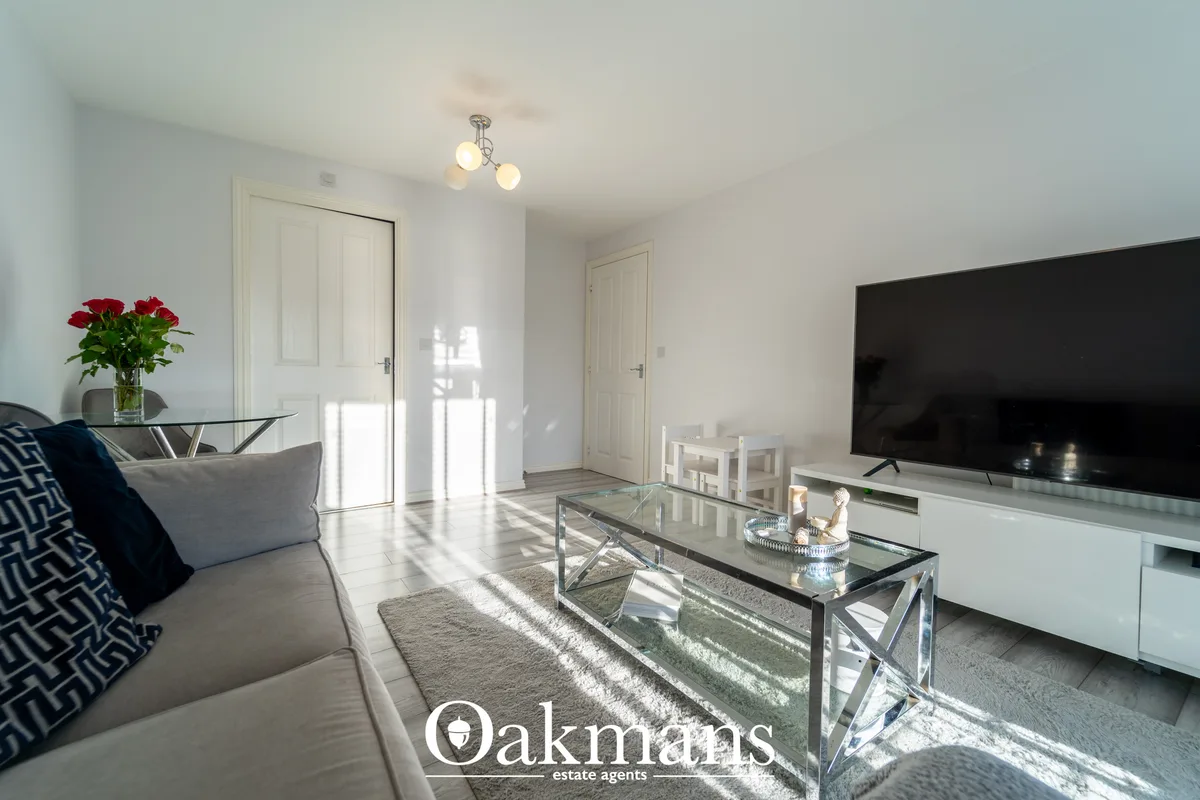 Bright living room with grey sofa, glass coffee table, wall-mounted TV, red flowers on side table, and French doors open