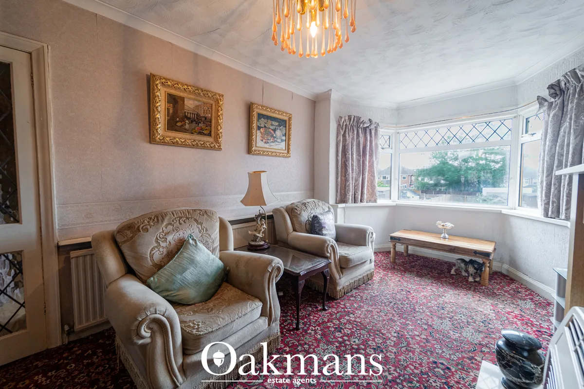 Elegant living room with beige upholstered armchairs, ornate gold-framed artwork, bay windows with patterned curtains, b