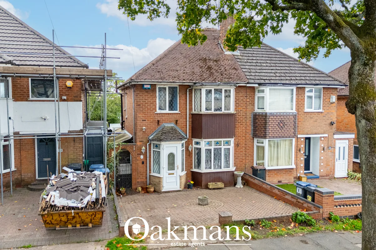Red brick semi-detached house with pitched roof, bay windows, white front door, paved driveway and mature tree