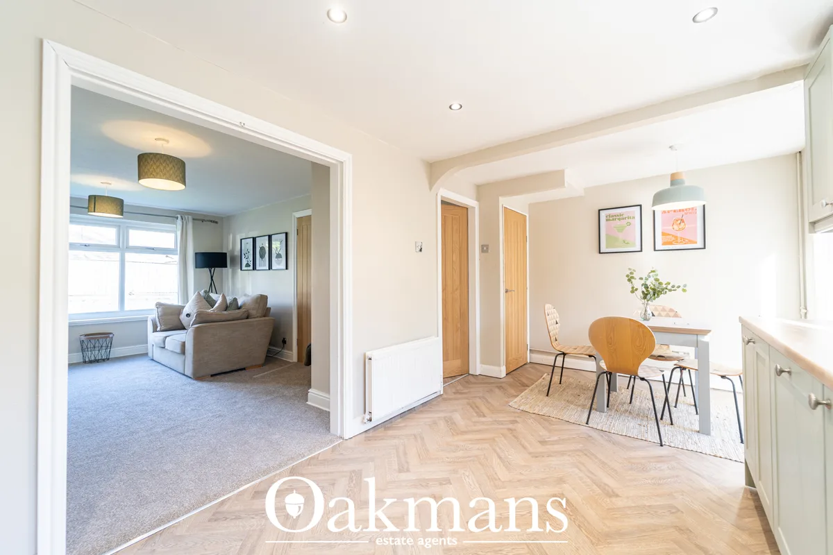 Bright entrance hallway with herringbone wood flooring, cream walls, open doorway to living room with carpet and armchai