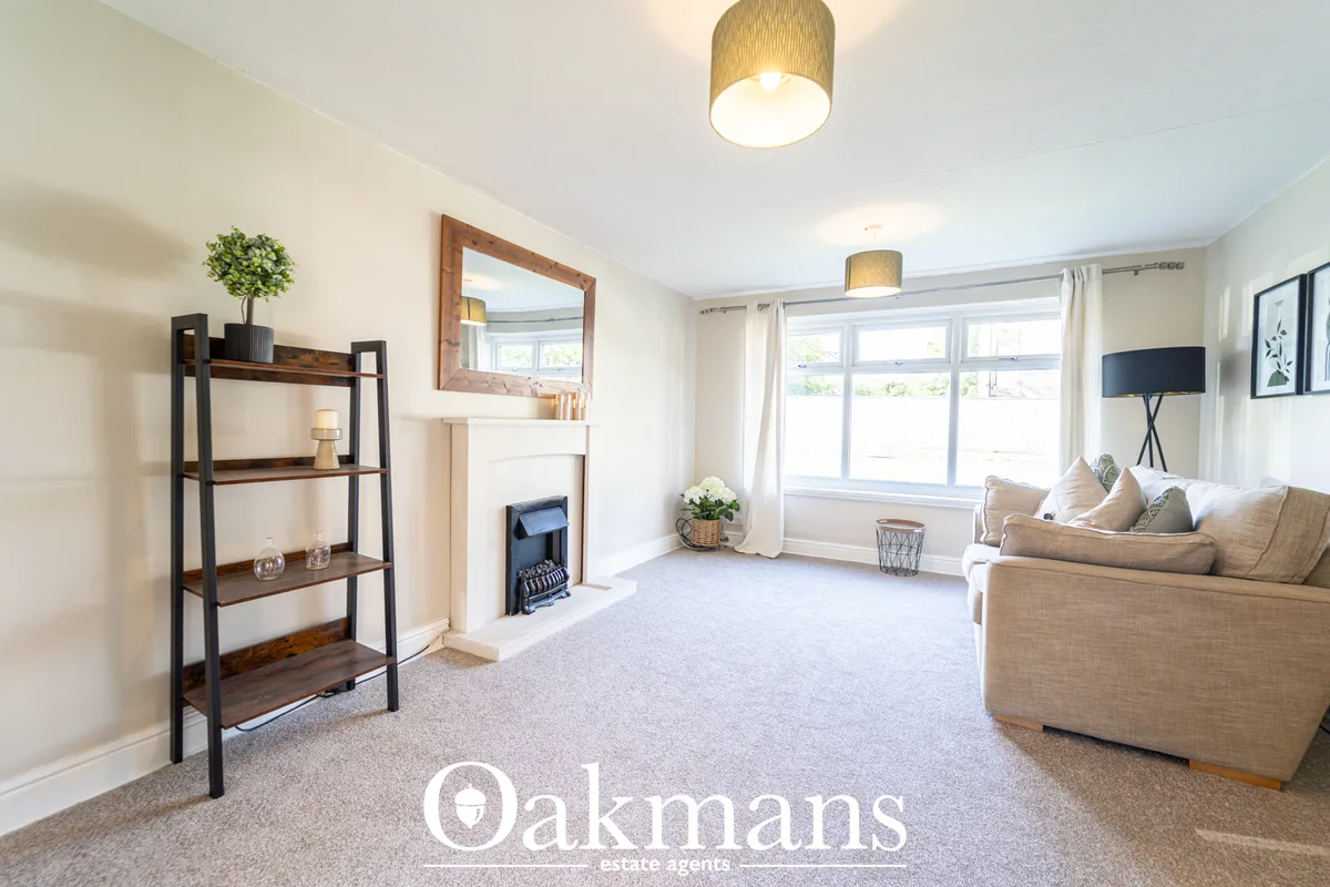 Bright living room with beige carpet, cream sofa, wooden shelving unit, fireplace, large windows with white curtains, ce