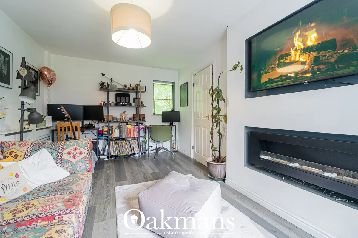 Open-plan living room with colourful patterned sofa, fireplace, desk area with dual monitors, shelving, indoor plants an