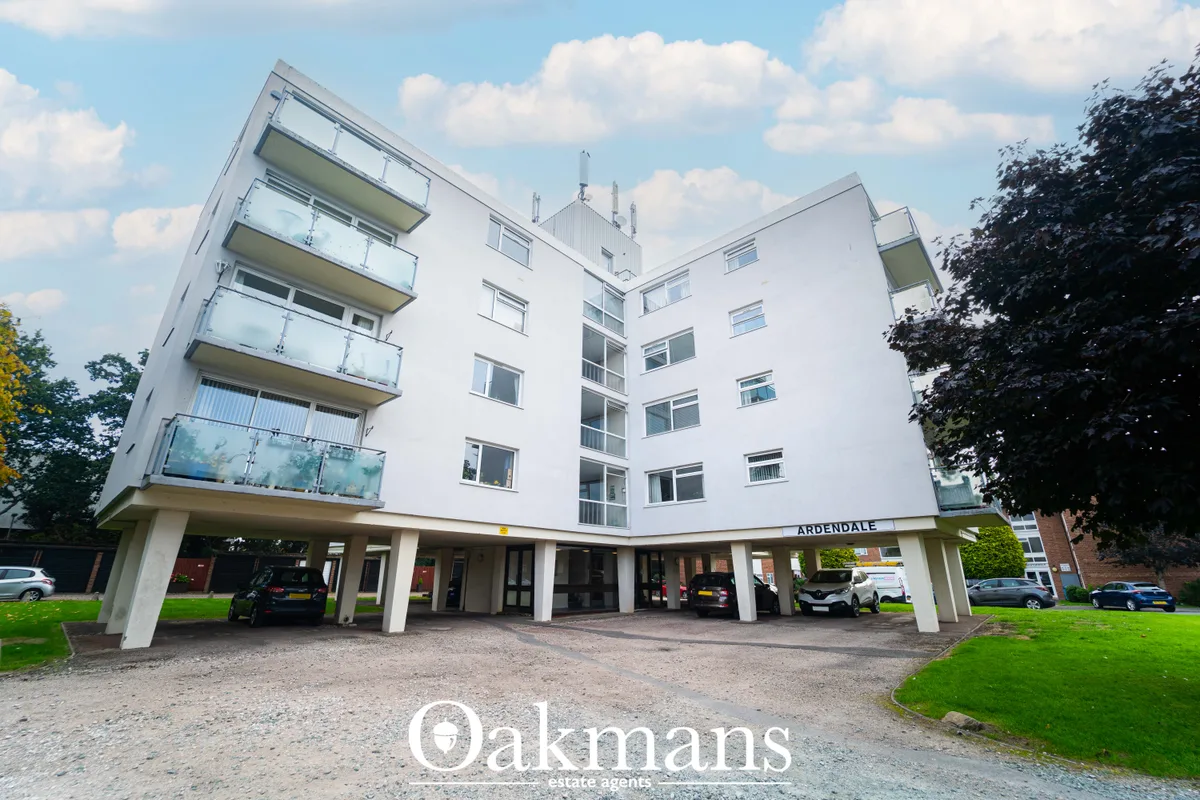 Modern multi-storey apartment building with white facade, glass balconies, ground-floor parking, surrounded by green gro