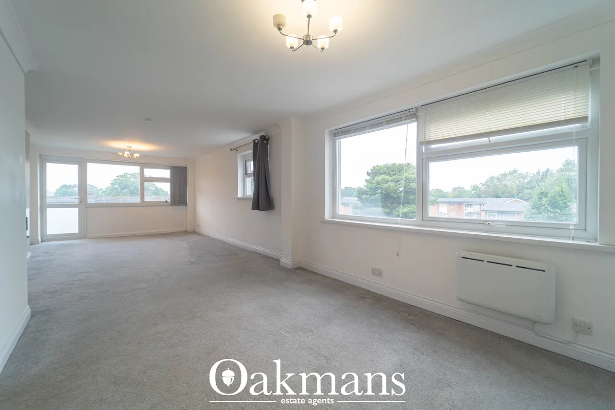 Spacious living room with multiple windows, beige carpet, ceiling light fixture, and views of surrounding greenery and r