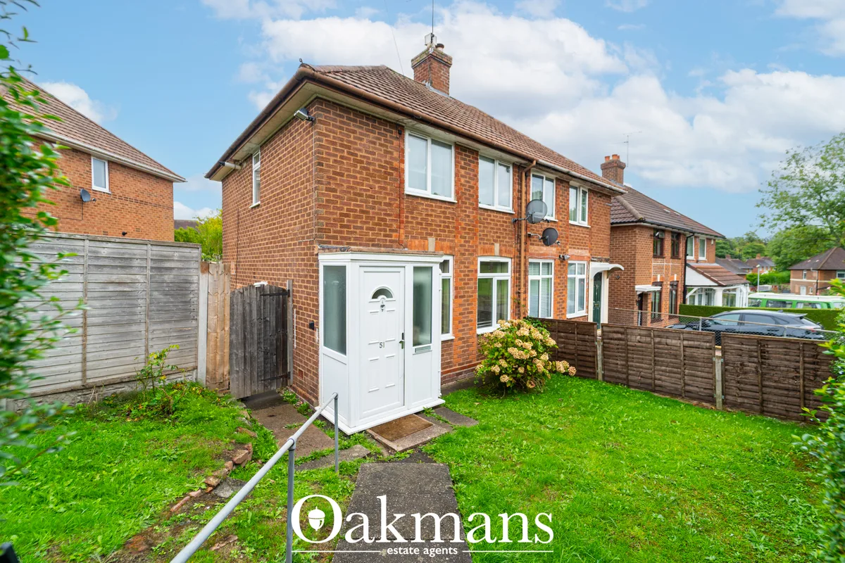 Red brick semi-detached house with white door porch, manicured lawn, wooden fence and flowering shrub