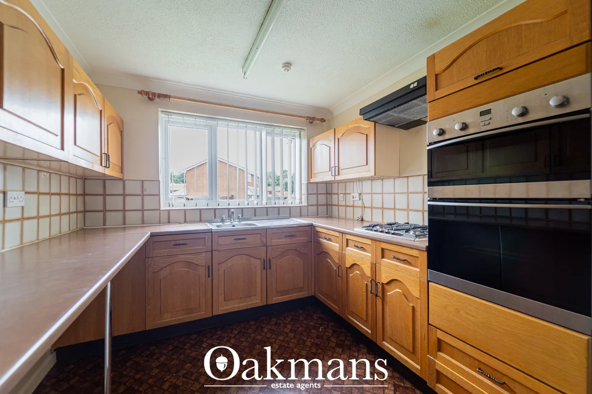 Fitted kitchen with honey wood units, beige tile splashback, stainless steel oven and hob, sink beneath window with vert