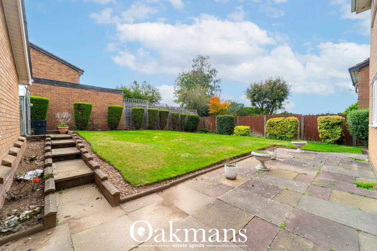 Large rectangular garden with manicured lawn, paved terrace, wooden fence, established hedges and trees, brick house vis