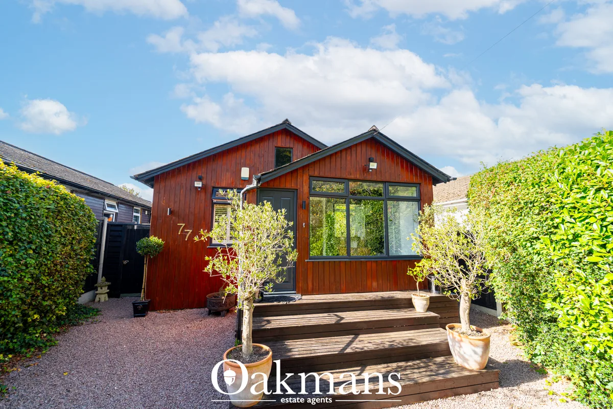 Modern red-brown timber-clad bungalow with dark roof, large glazed windows, wooden deck entrance, manicured hedges and g