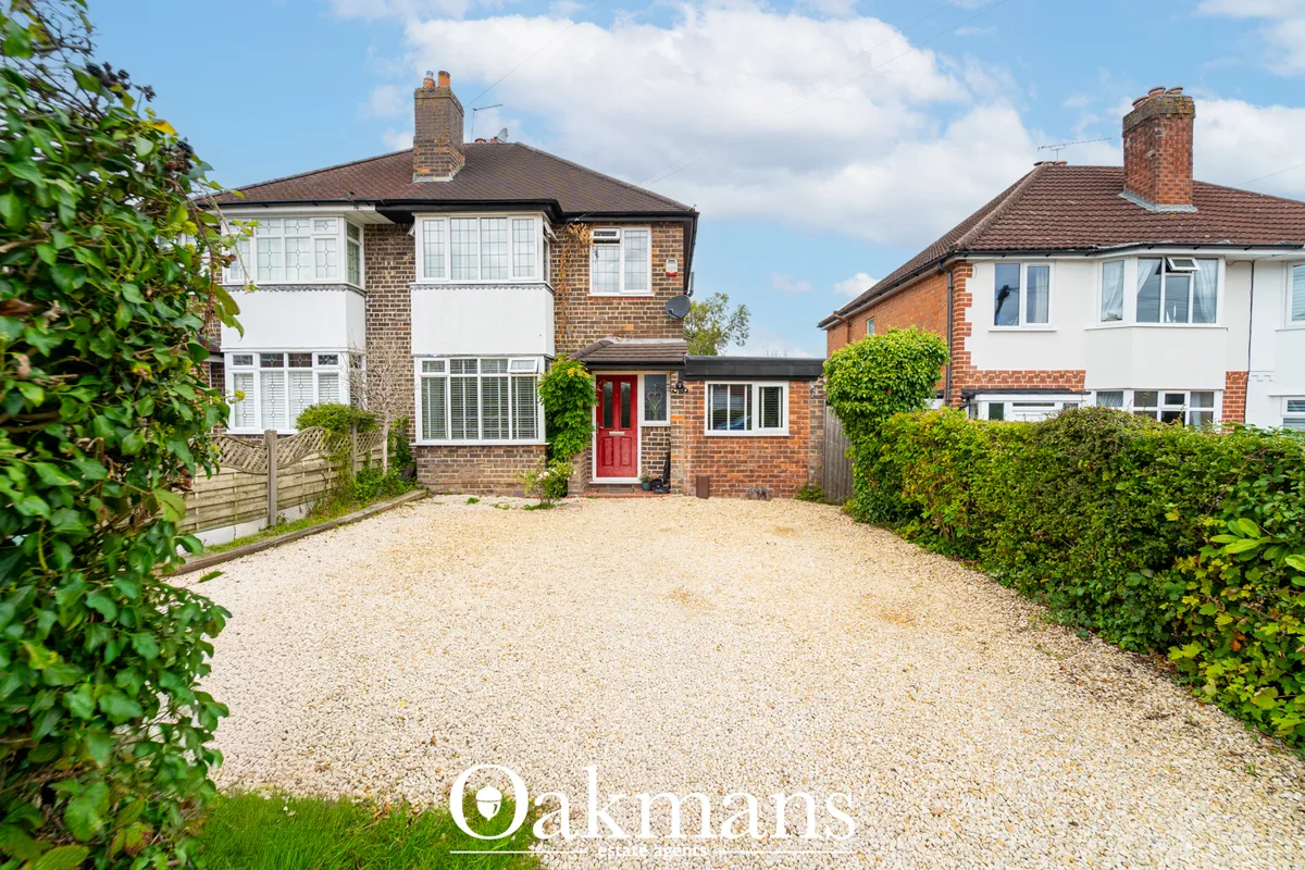 Semi-detached brick house with white-framed windows, red front door, gravel driveway, and green hedging borders
