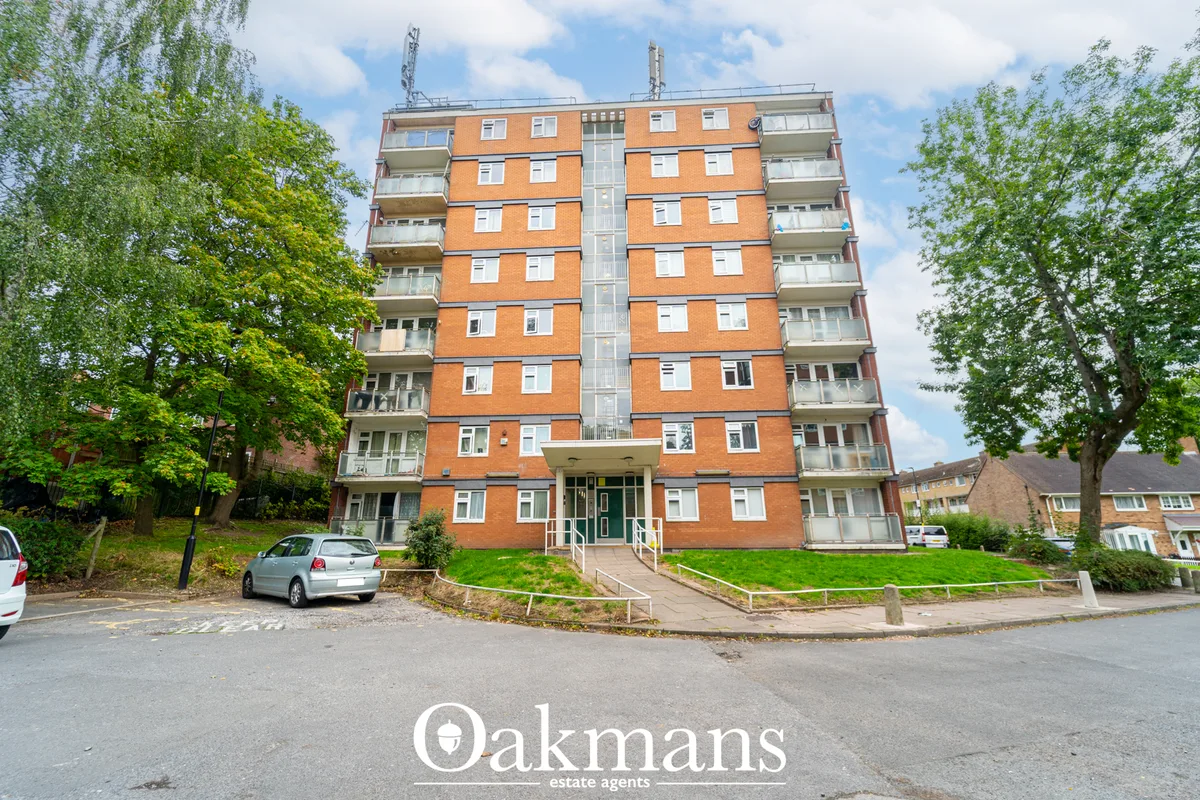 Orange-brick residential apartment building with eight storeys, white balconies, central entrance, surrounding trees and