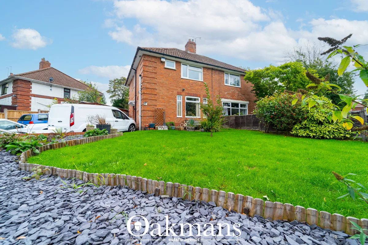 Red brick semi-detached house with manicured front lawn, gravel border, wooden fence, and white van parked on driveway