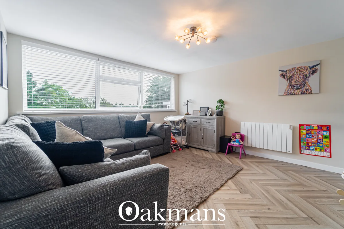 Spacious living room with grey corner sofa, neutral decor, wooden flooring, large window with blinds, and children's pla
