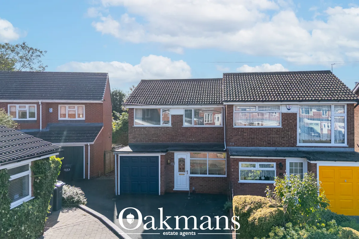 Red brick semi-detached houses with dark tile roofs, driveways, and garages. Clear blue sky with white clouds