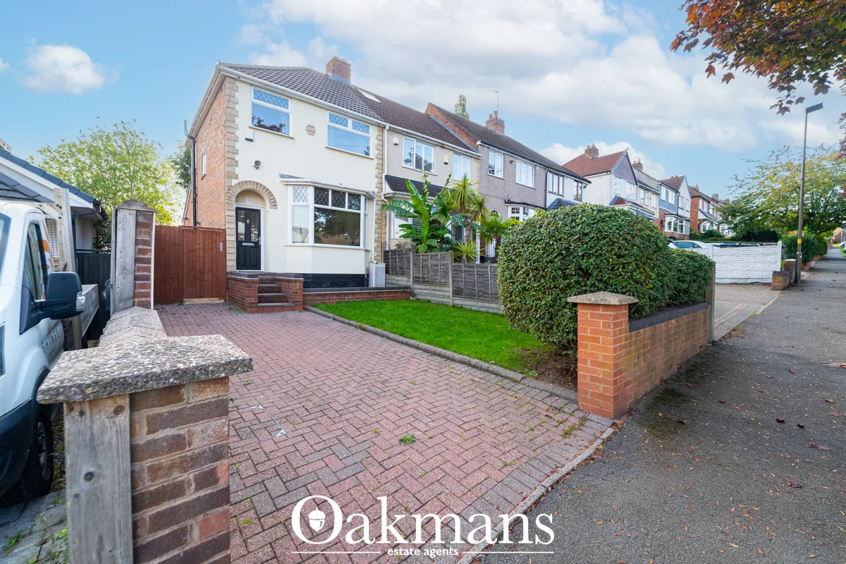 Semi-detached brick house with white rendered upper storey, pitched roof, brick driveway, hedged forecourt and neighbour
