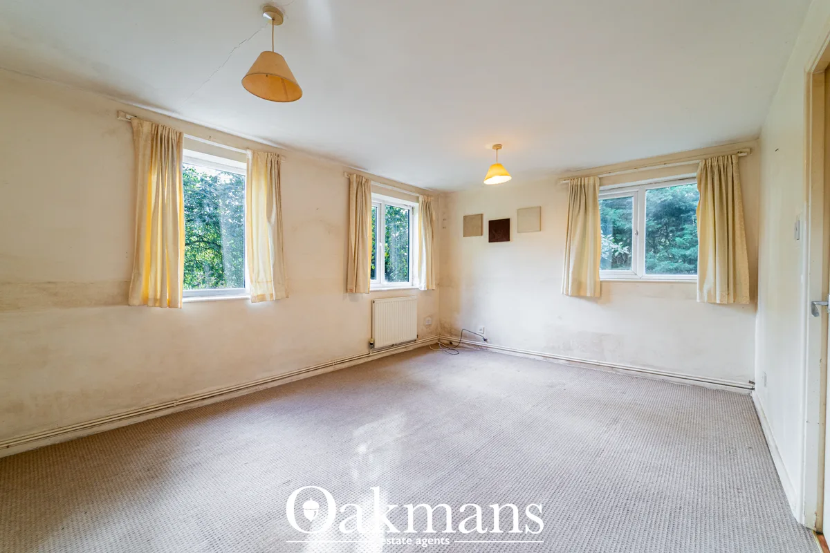 Spacious living room with cream walls, carpeted floor, three windows with pale gold curtains, two pendant lights, radiat