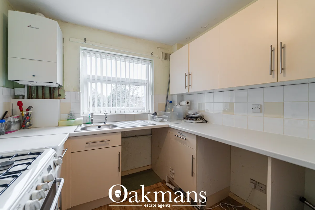 Fitted kitchen with beige and cream units, white sink, gas cooker, boiler, tiled splashback and window with vertical bli