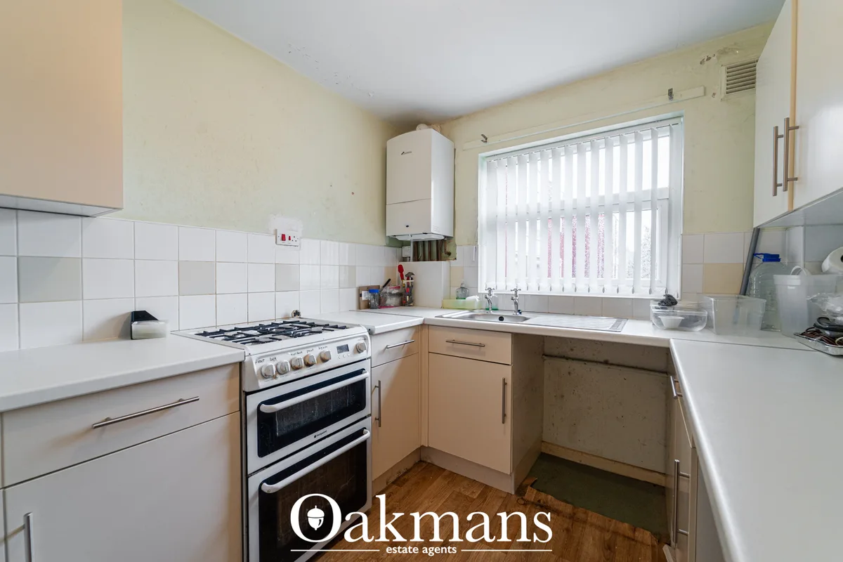 Fitted kitchen with cream and beige units, white tile splashback, gas cooker, sink under window with vertical blinds, wa