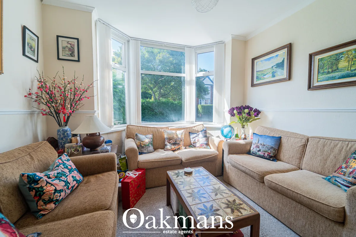 Bright living room with bay windows, beige upholstered sofas, decorative pillows, framed landscape artwork, and ornament