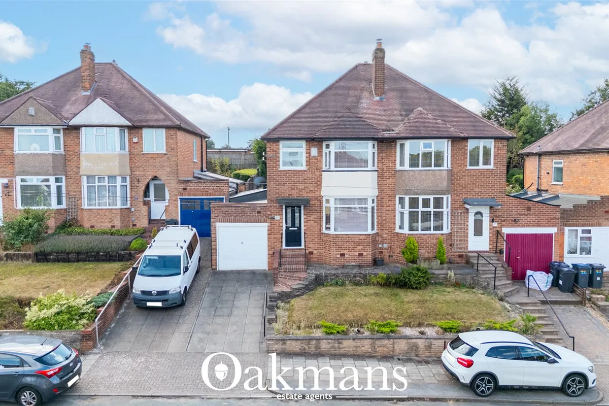 Suburban street view showing red brick detached houses with pitched roofs, driveways, parked cars and manicured gardens