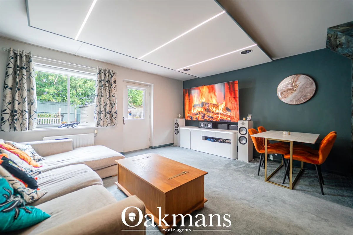 Open-plan living and dining space with beige sofa, wooden coffee table, wall-mounted TV displaying fireplace, orange vel