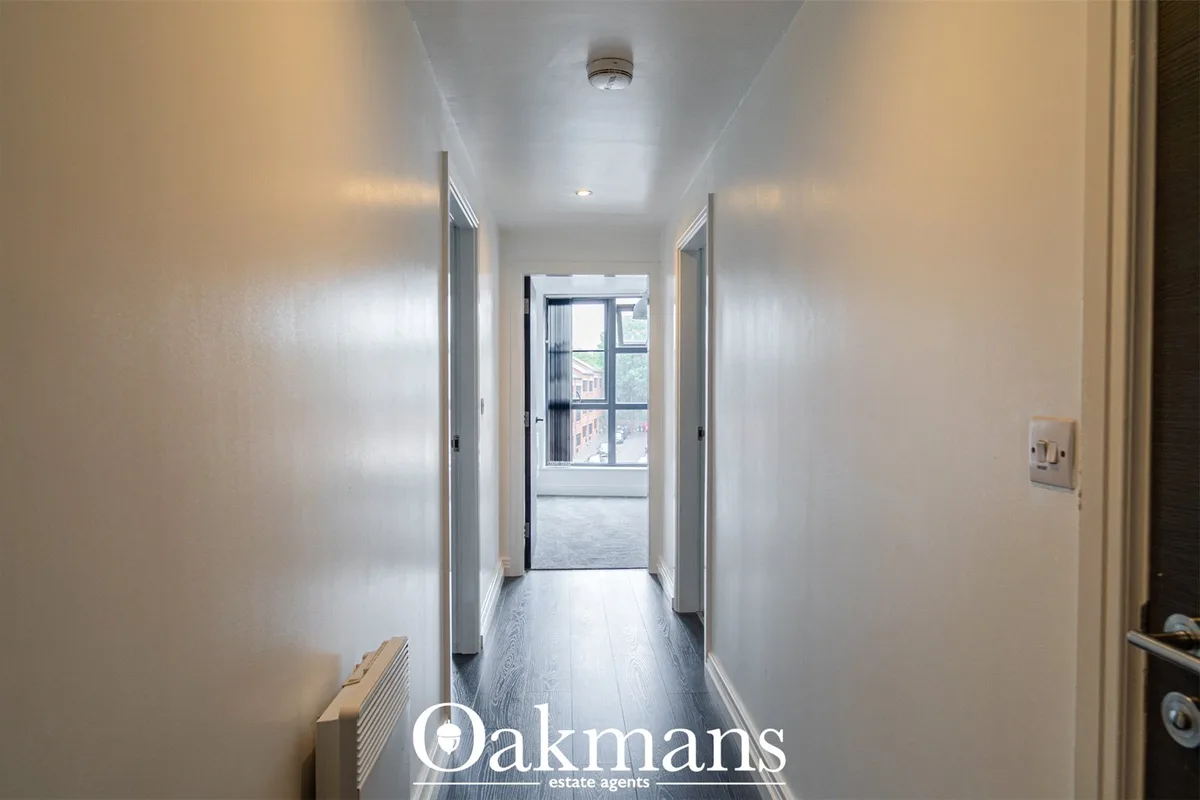 Bright hallway with white walls, grey wood flooring, ceiling light fixture and open door to bright room with windows