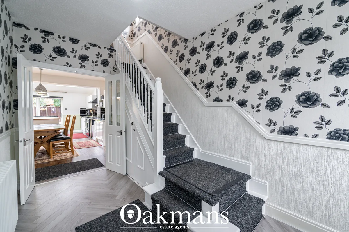 Bright hallway with staircase, black floral wallpaper, white panelling, wooden flooring and open doorway to dining area