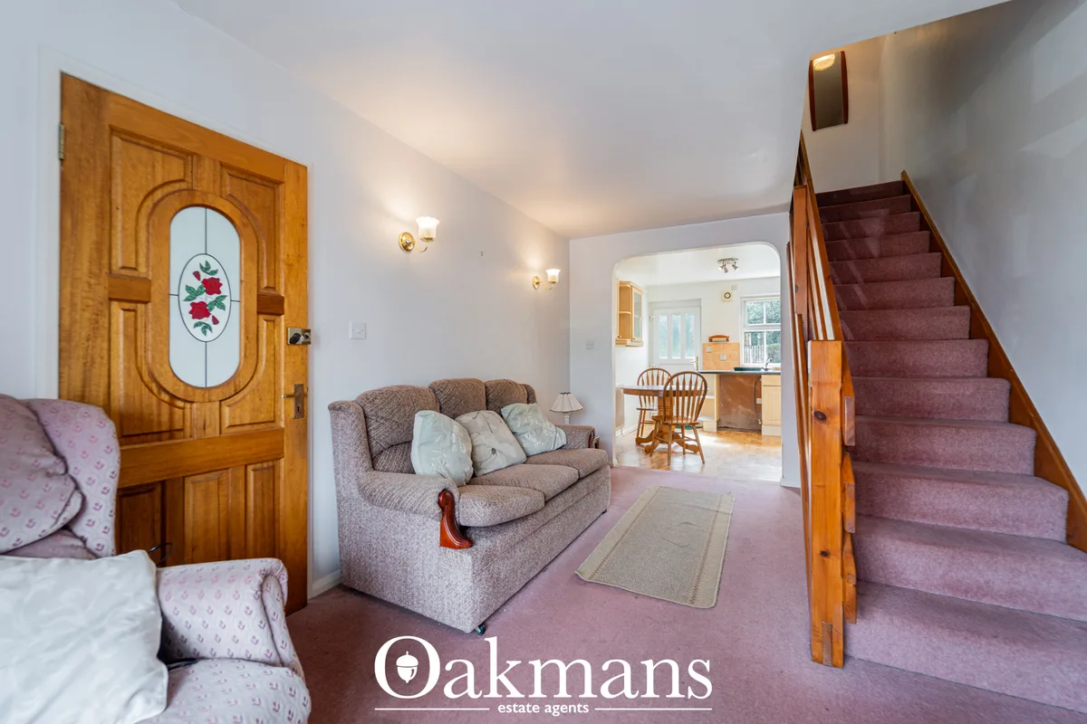 Entrance hallway with ornate wooden door, staircase with burgundy carpet, beige sofa, and views through to kitchen and d
