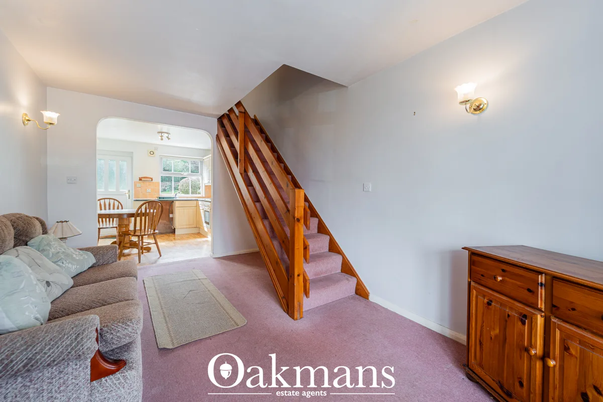 Bright hallway with wooden staircase, pink carpet, white walls, wooden dresser and access to kitchen