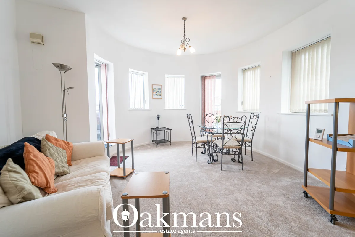 Bright open-plan living space with cream sofa, dining table with metal chairs, multiple windows with vertical blinds, pe
