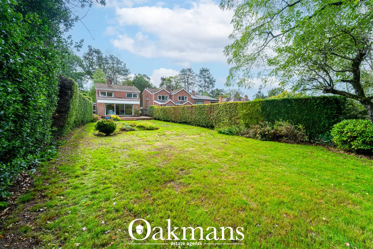 Rear garden with expansive lawn, mature trees, brick houses and manicured hedges under blue sky