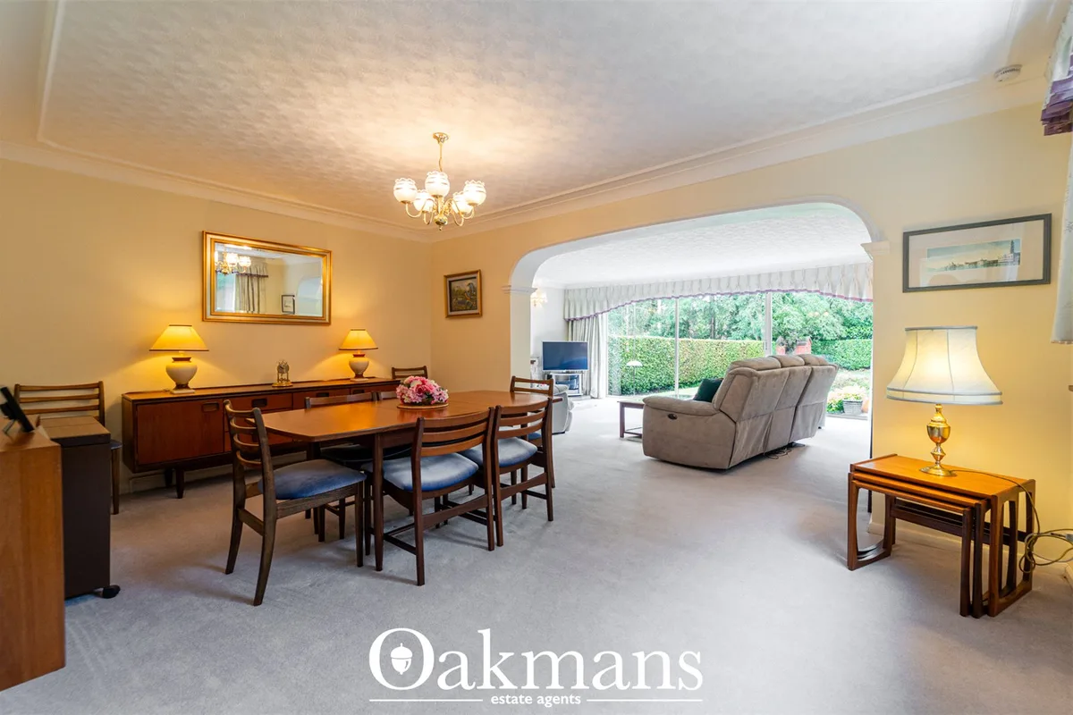Spacious living room with cream walls, wooden dining table with four chairs, beige reclining sofa, and French doors open