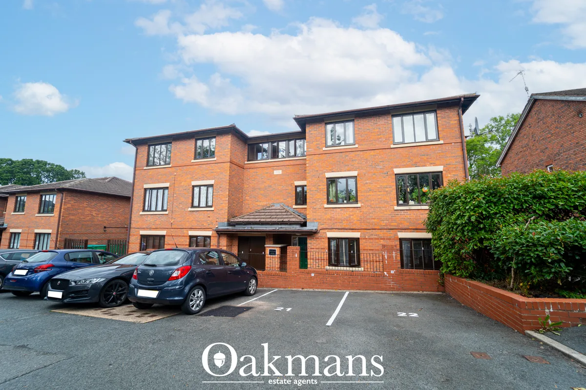 Modern red-brick apartment building with multi-level design, parked cars, and manicured hedging