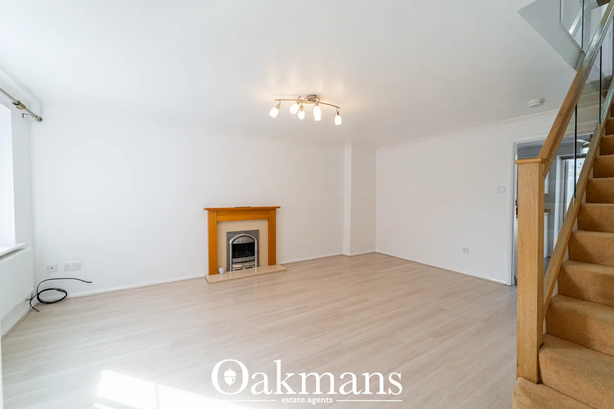 Spacious living room with wooden fireplace surround, light wood flooring, ceiling lights, stairs to upper level, and whi