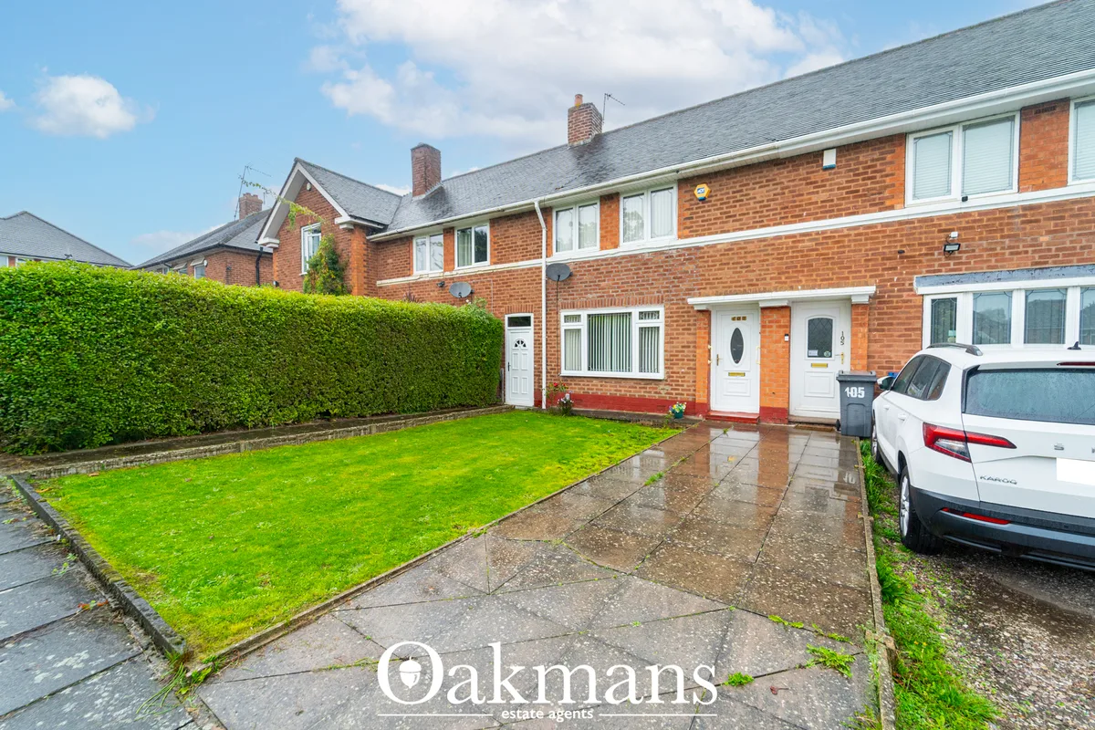 Red brick terraced house with front garden, driveway with white car, green hedge boundary, and grey slate roof