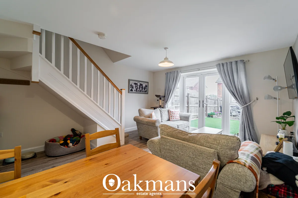Bright living room with beige upholstered seating, wooden dining table, staircase, grey curtains and French doors openin