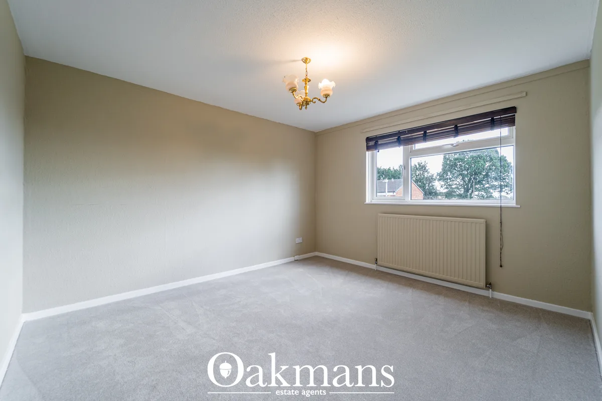 Empty bedroom with beige and white walls, carpet flooring, radiator, large window with tree views, and ceiling light fix